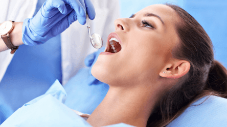 A dentist examines a female patient’s open mouth with a dental mirror.